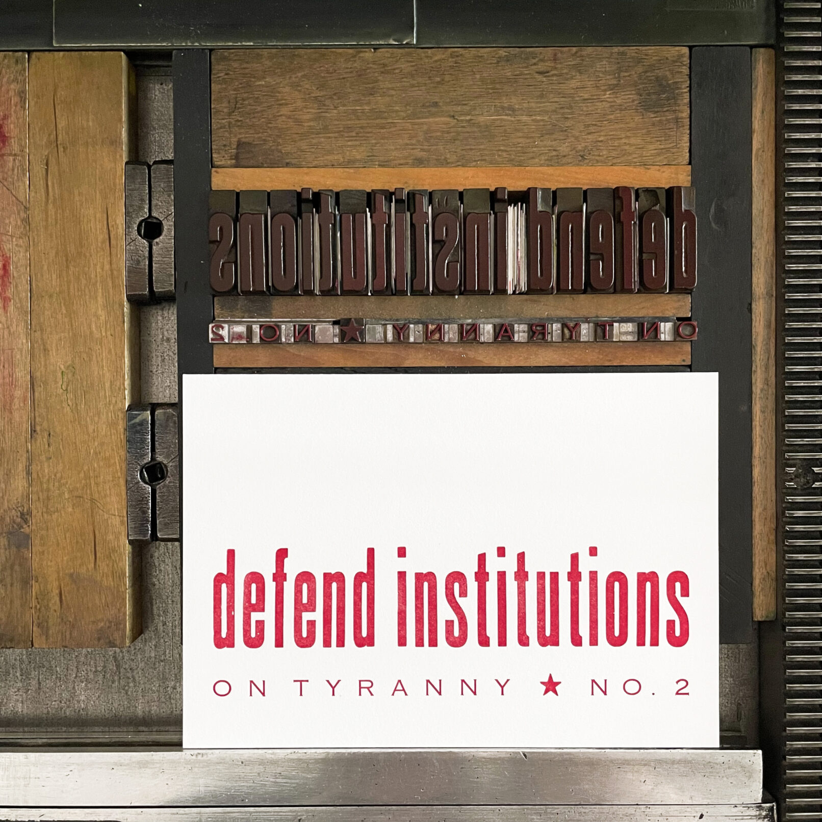 Letterpress print on Vandercook press bed. Printed in red ink on white paper, the text reads "Defend Institutions On Tyranny No. 2."
